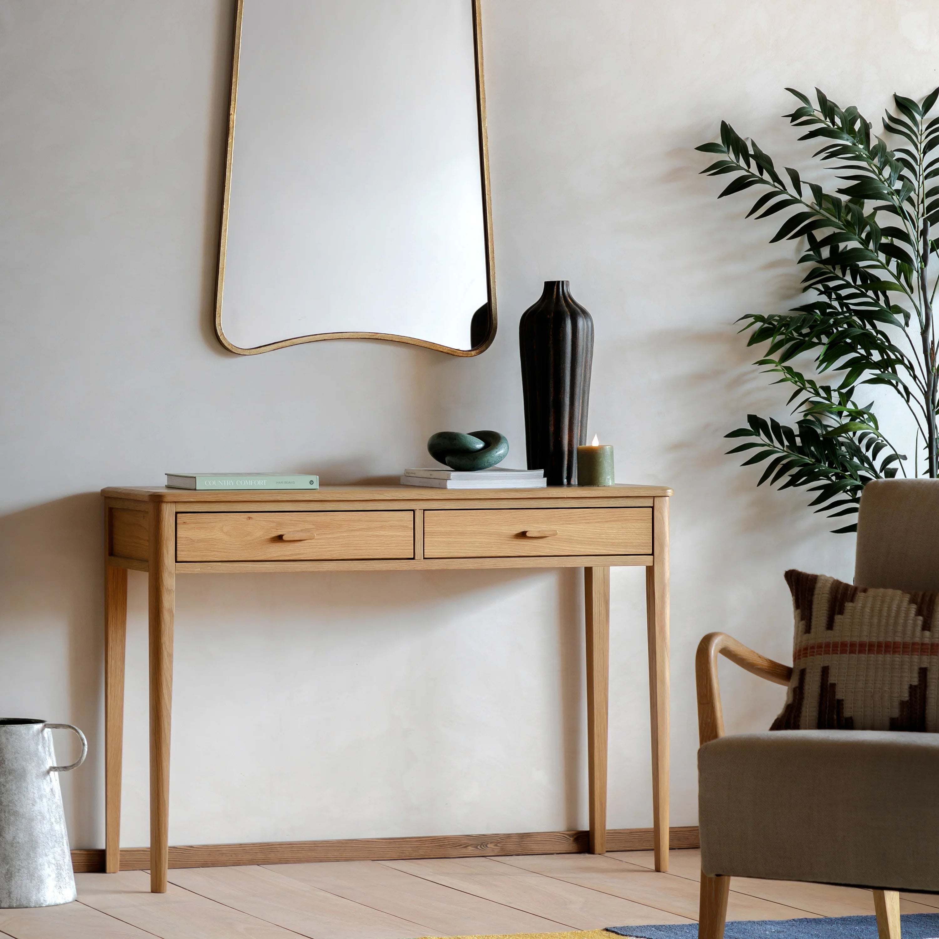 A light natural wood console table with two drawers and slim legs, styled in a minimalist room with a large wavy mirror, a black vase, books, and a potted plant.
