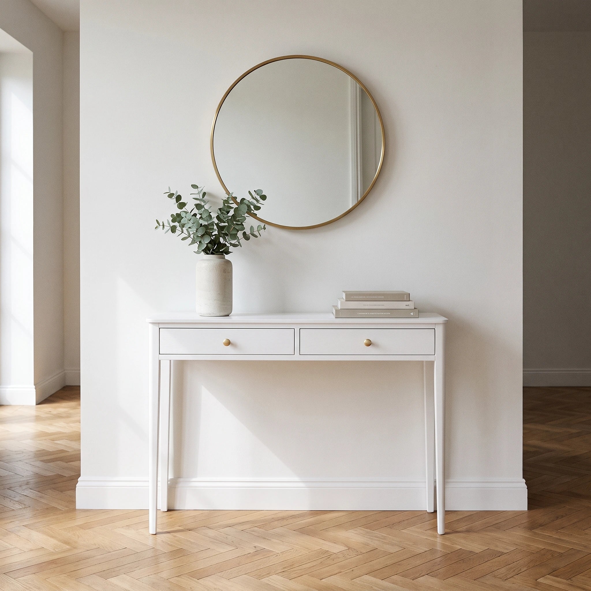A white console table with two drawers and small gold knobs, styled with a vase of greenery and books, beneath a round gold-framed mirror on a white wall over a herringbone wood floor.